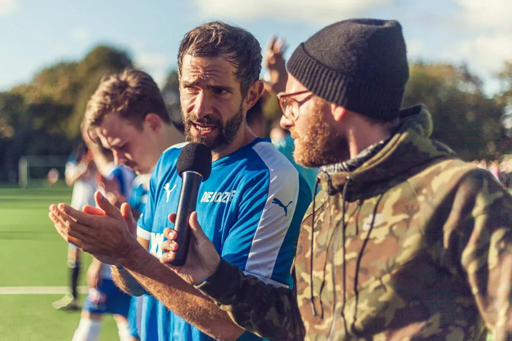 Mann mit Mütze hält einem Fußballer im blauen Trikot ein Mikrofon hin, auf einem sonnigen Fußballplatz.
