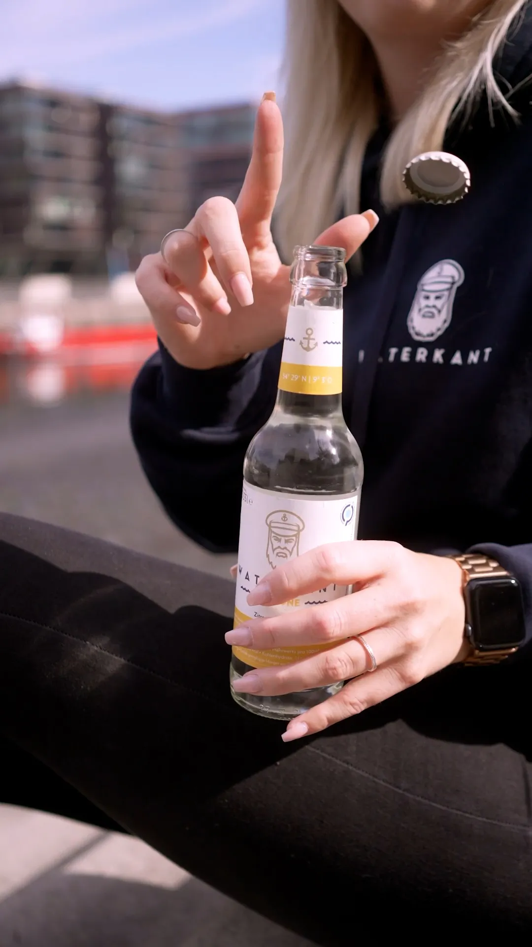 Person im schwarzen Waterkant-Hoodie öffnet eine Glasflasche im Sonnenlicht; der Kronkorken fliegt durch die Luft.