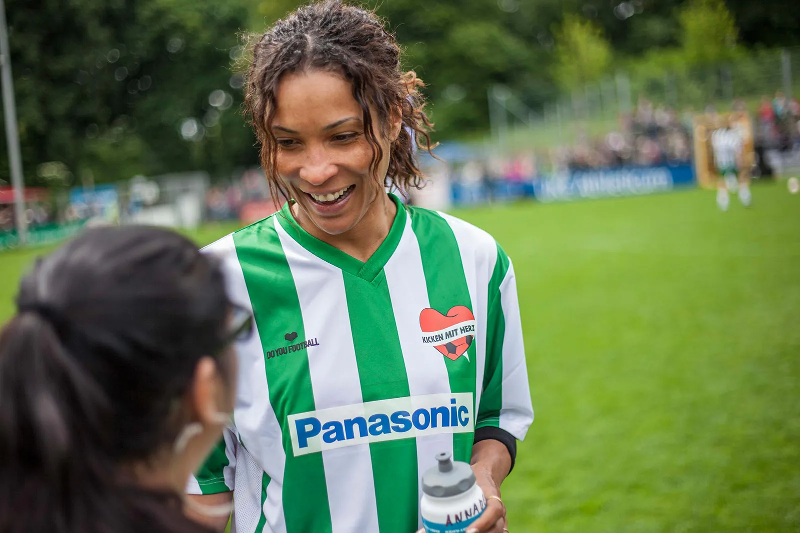 Lächelnde Person im grün-weißen Kicken-mit-Herz-Trikot mit Trinkflasche im Gespräch am Spielfeldrand.