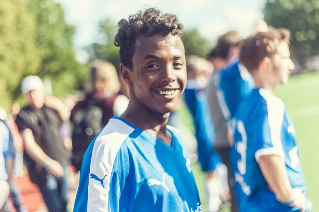 Lächelnder Fußballspieler im blauen Trikot am sonnigen Spielfeldrand, weitere Spieler im Hintergrund.
