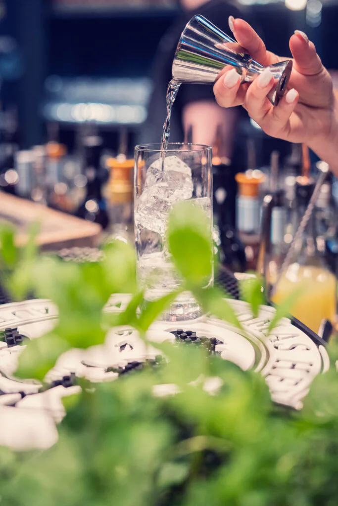 Eine Hand gießt an einer Bar Flüssigkeit aus einem Metallmessbecher in ein hohes Glas mit Eis; vorn Minze.