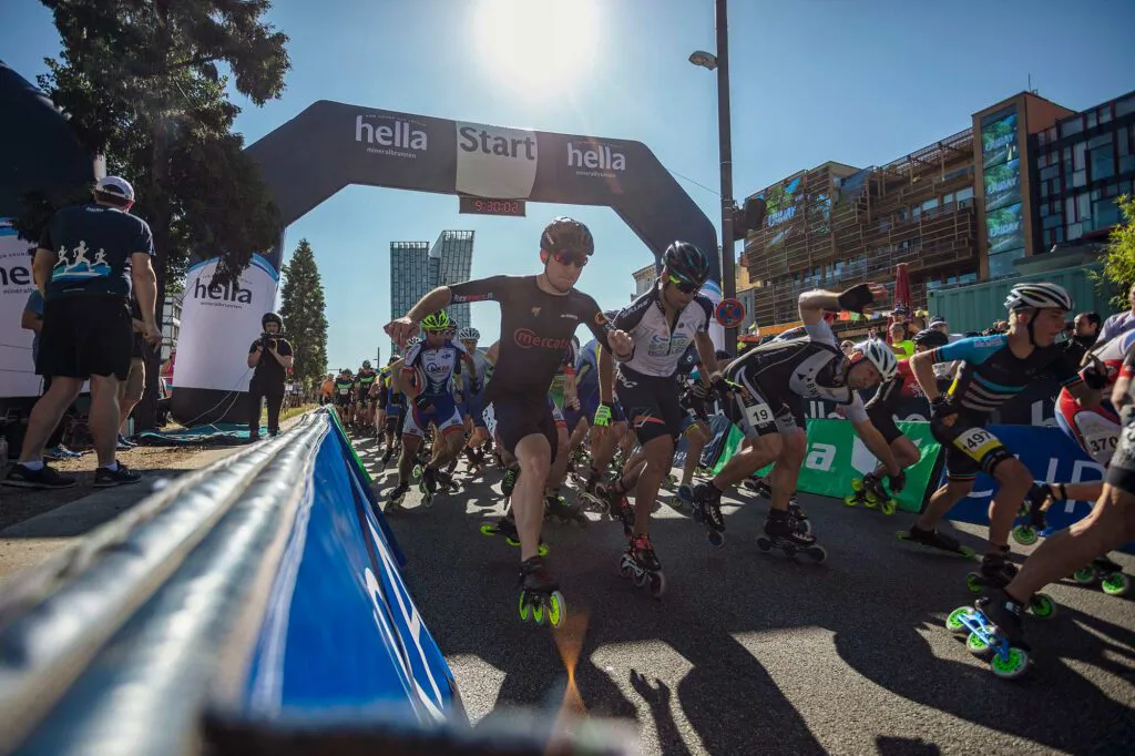 Inline-Skater starten bei starkem Sonnenlicht unter einem hella-Startbogen auf einer abgesperrten Straße.