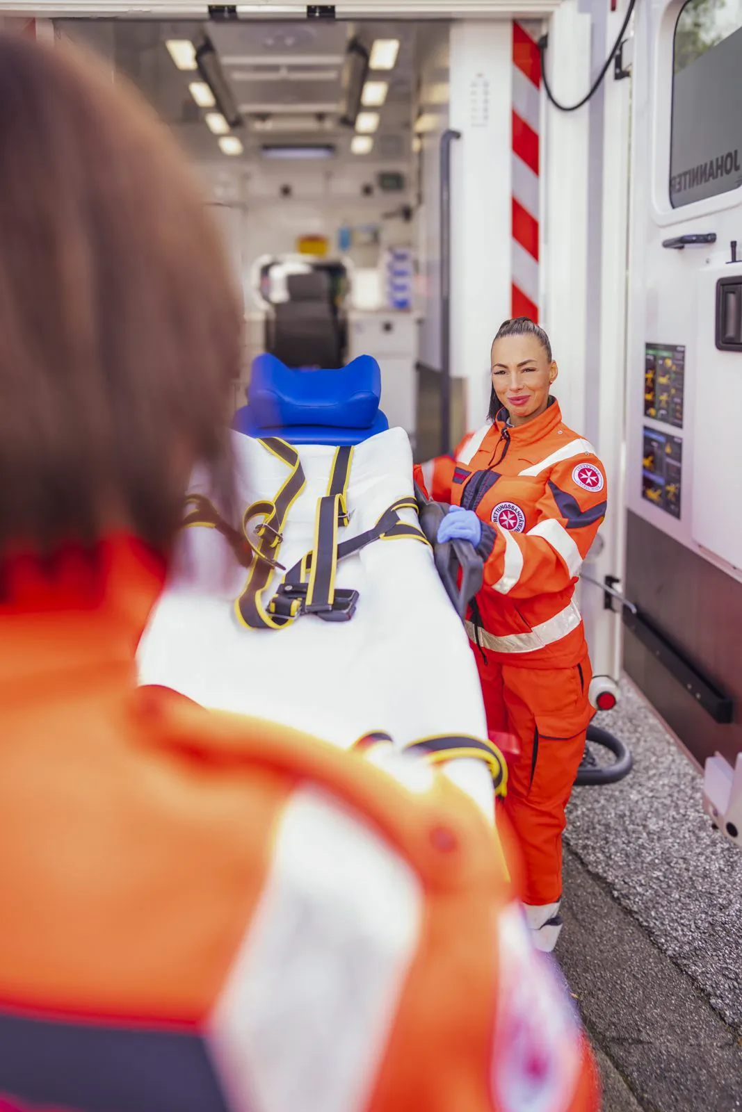 Zwei Johanniter-Rettungskräfte in orangefarbener Einsatzkleidung halten eine Trage am offenen Rettungswagen.