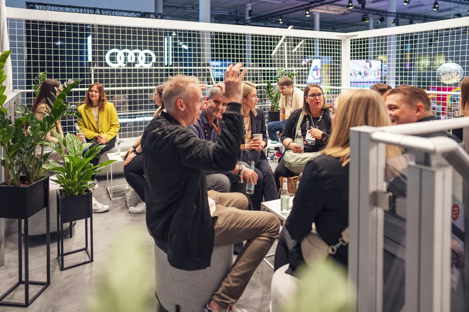 Besucher sitzen im hellen Awin-Messestand zwischen Pflanzen; ein Mann hebt im Gespräch die Hand.
