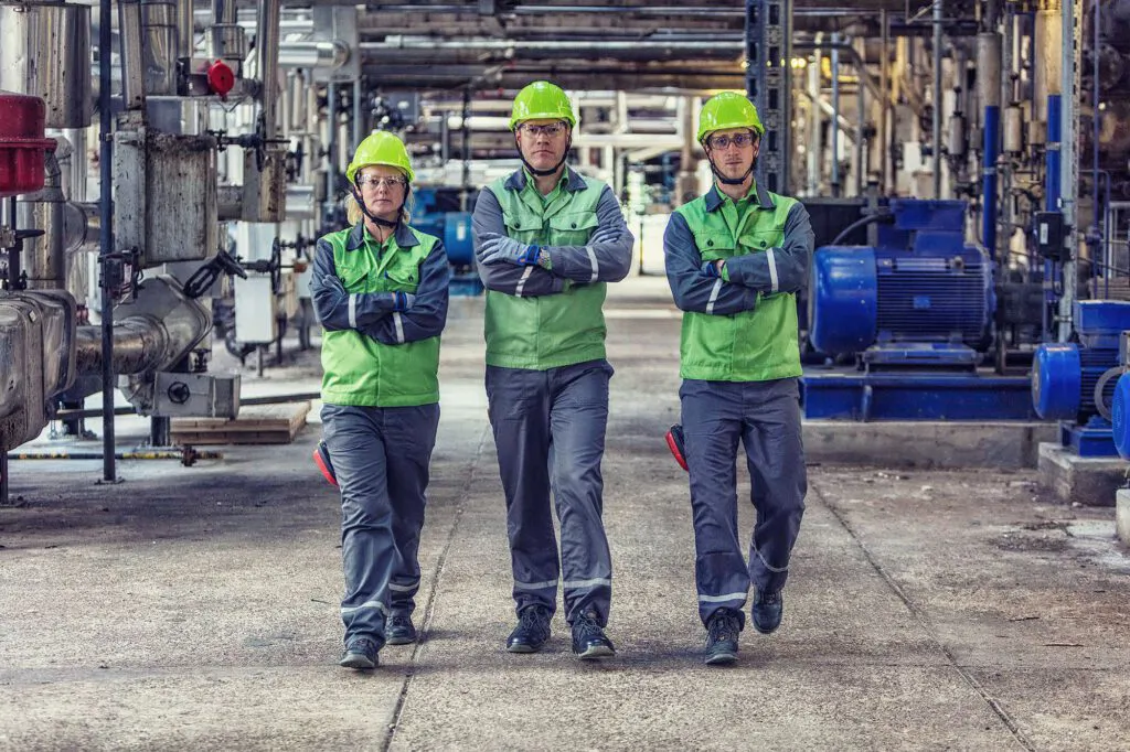 Drei Beschäftigte mit grünen Helmen und verschränkten Armen zwischen Rohren und Pumpen in der Raffinerie Heide.
