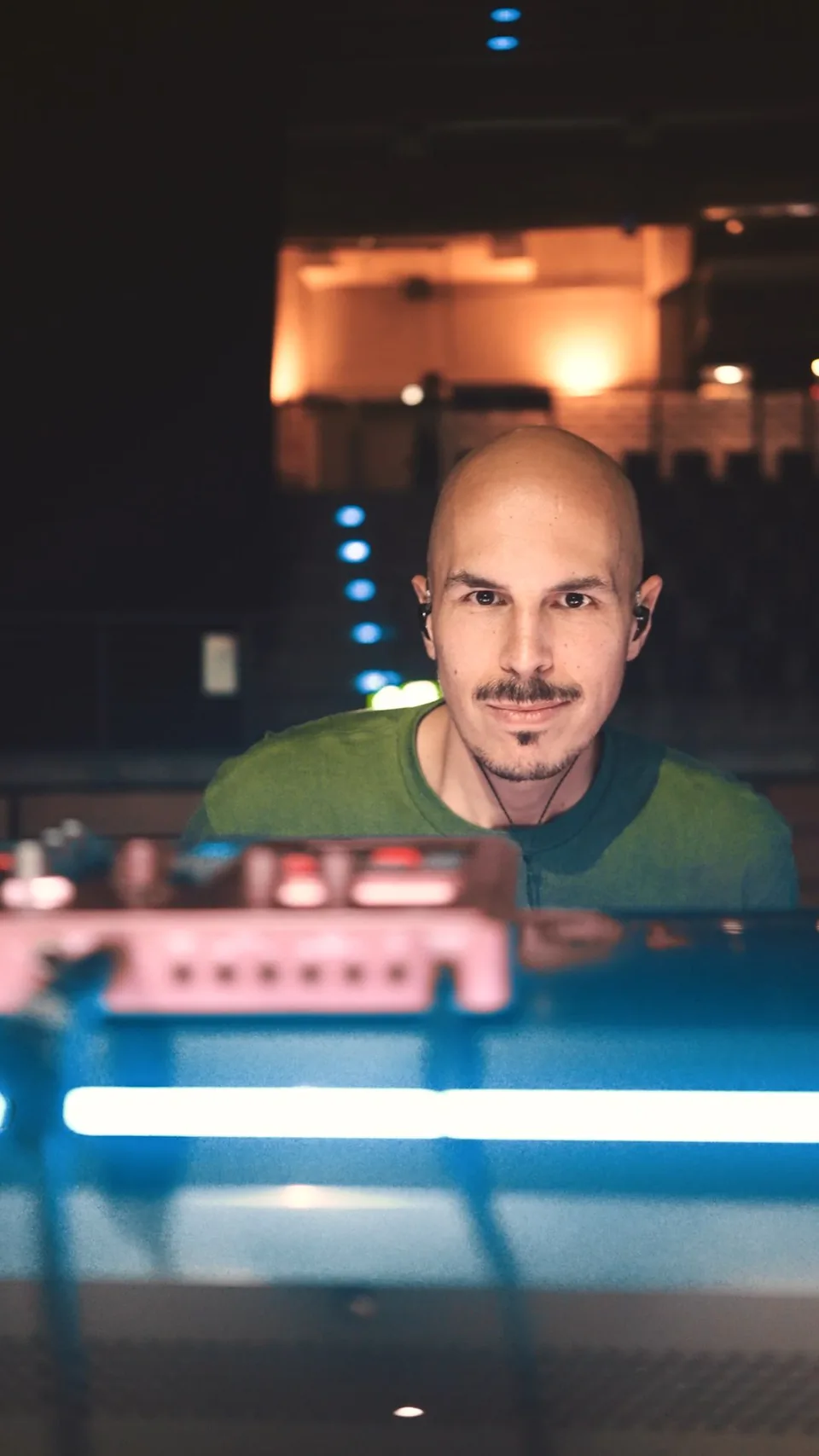 Mann mit In-Ear-Kopfhörern hinter einem Mischpult, im Vordergrund blaues Licht, dahinter ein warm beleuchteter Saal.