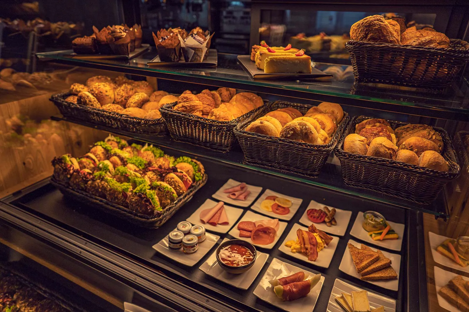 Warm beleuchtete Buffetvitrine mit Brötchen, belegten Broten, Kuchen und kleinen Tellern mit Aufschnitt und Käse.