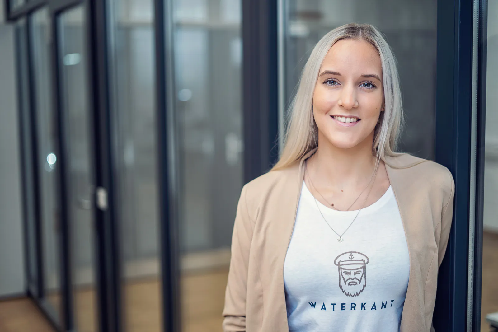 Lächelnde blonde Frau im beigen Blazer und Waterkant-Shirt vor einer Glaswand bei weichem Tageslicht.