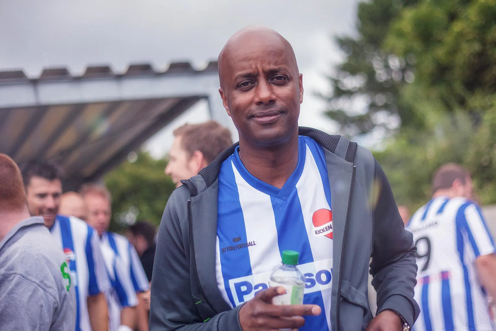 Mann im blau-weiß gestreiften Fußballtrikot hält eine Wasserflasche; weitere Spieler stehen draußen hinter ihm.