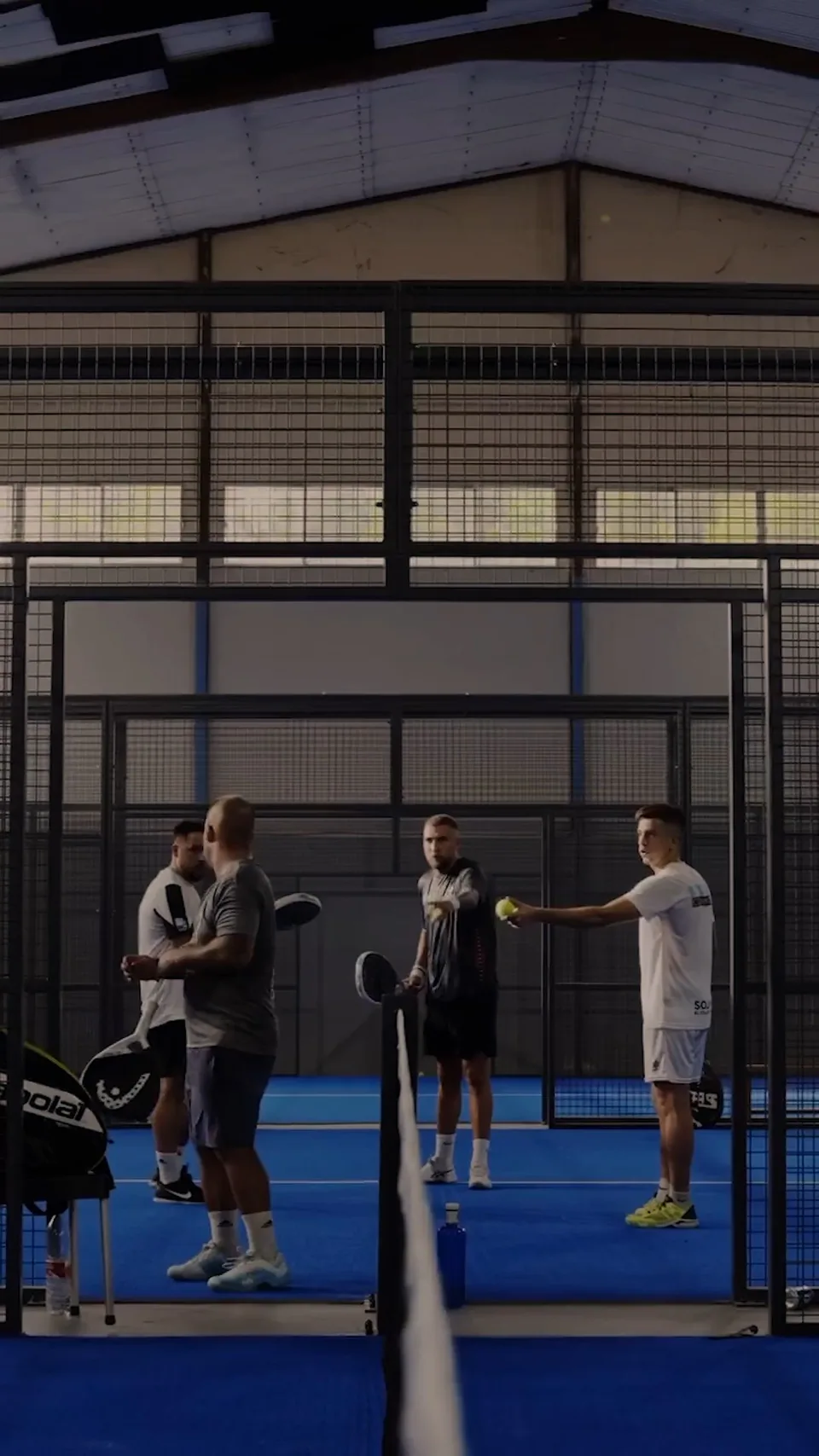 Spieler mit Padelschlägern stehen hinter einem Netz auf einem blauen Hallenplatz; einer hält einen gelben Ball.