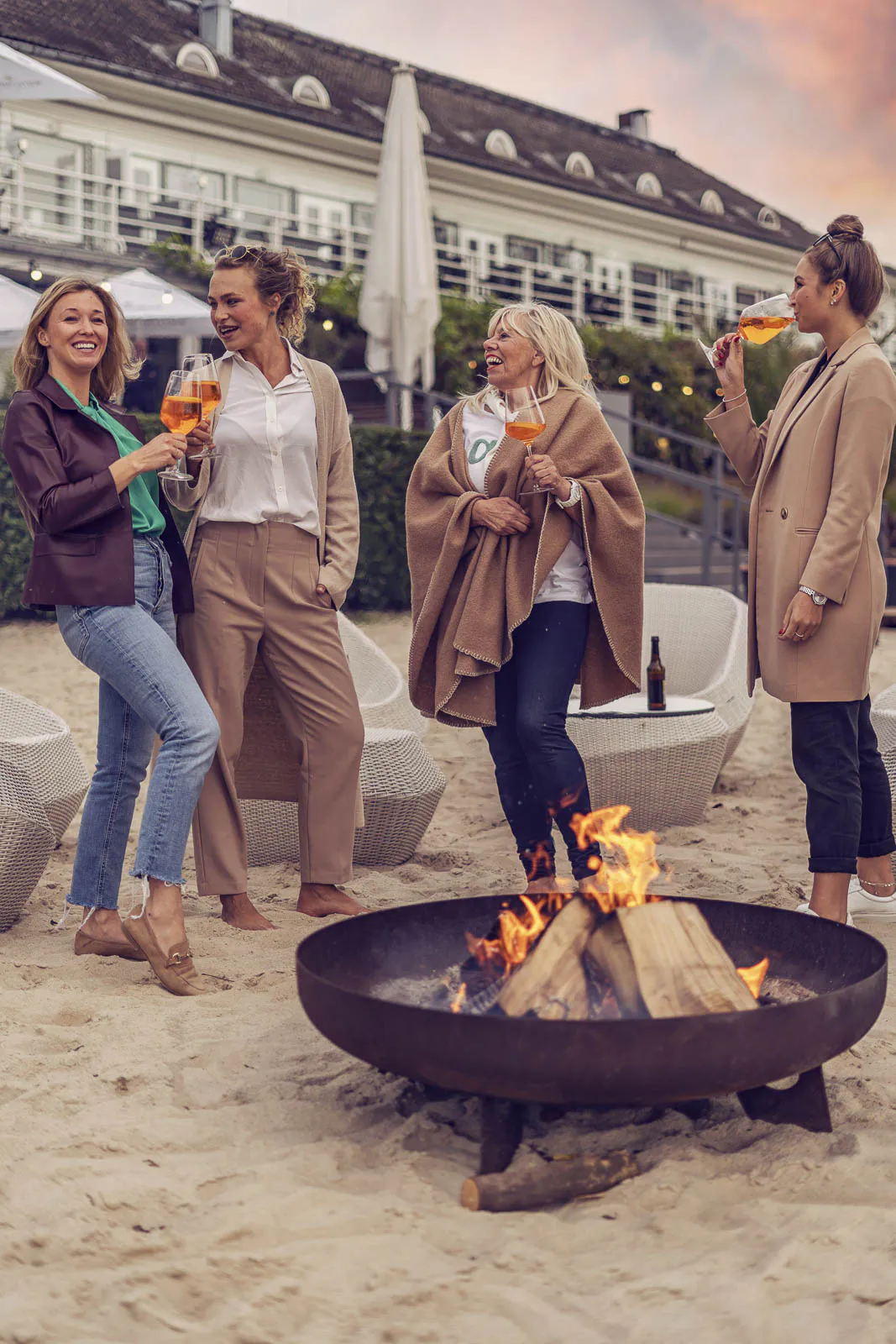 Vier Frauen mit orangefarbenen Drinks stehen im Sand eines Beachclubs um eine brennende Feuerschale im Abendlicht.