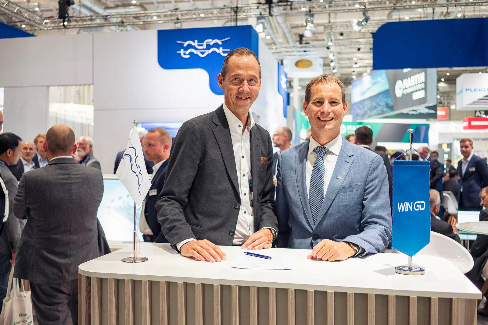 Zwei Männer in Anzügen stehen lächelnd hinter einem weißen Tresen am hell beleuchteten Messestand von Alfa Laval.