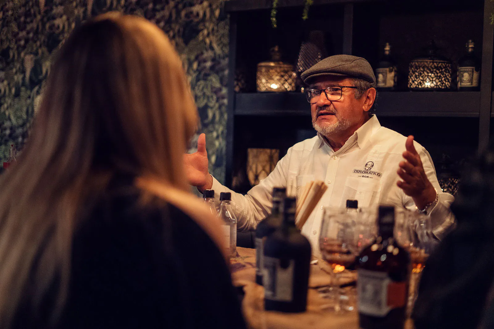Mann mit Hut gestikuliert hinter einer Bar mit Flaschen und Gläsern; davor Gäste im warmen Licht.
