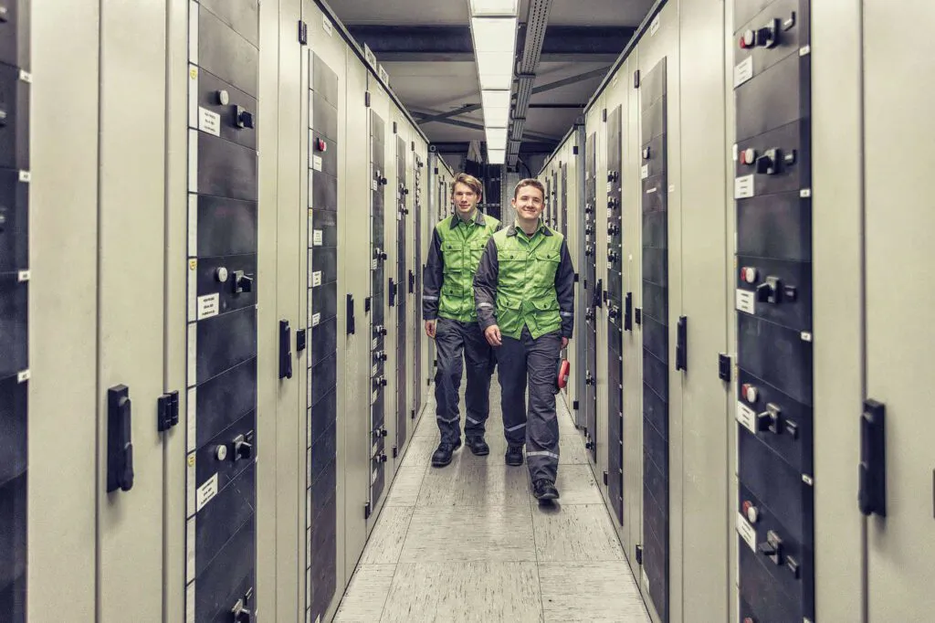 Zwei Personen in grüner Arbeitskleidung gehen durch einen beleuchteten Gang zwischen Schaltschränken der Raffinerie Heide.