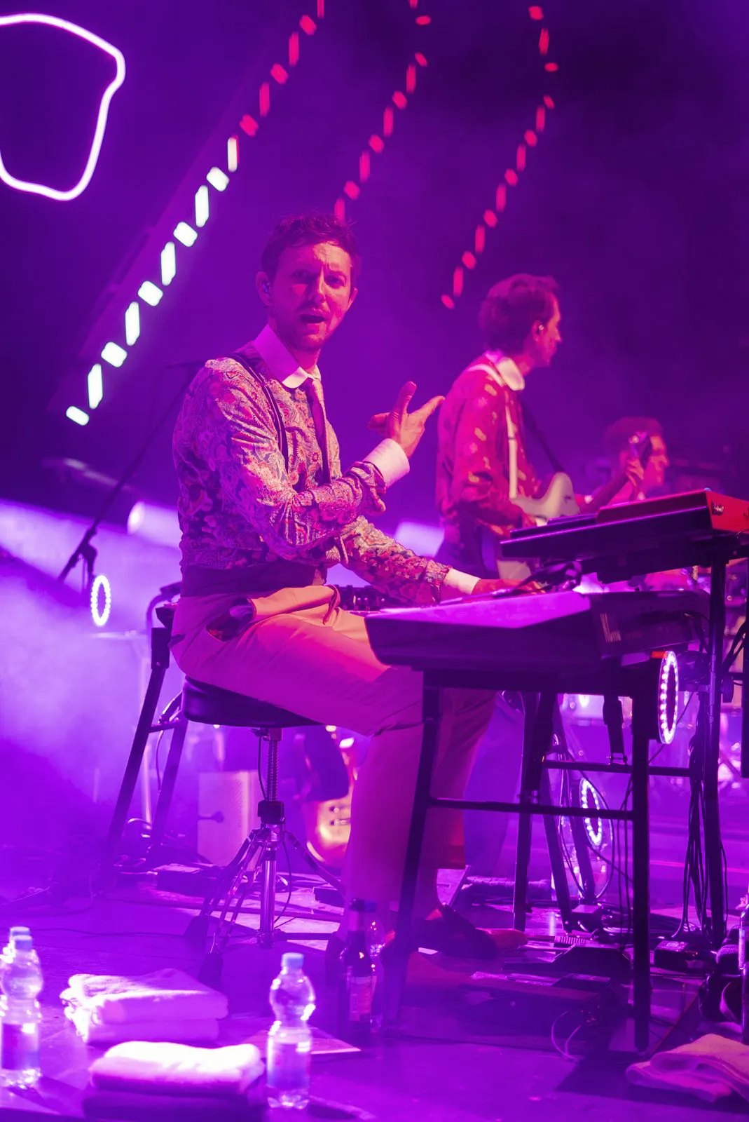 Keyboarder sitzt am E-Piano auf violett beleuchteter Konzertbühne, dahinter ein Gitarrist.