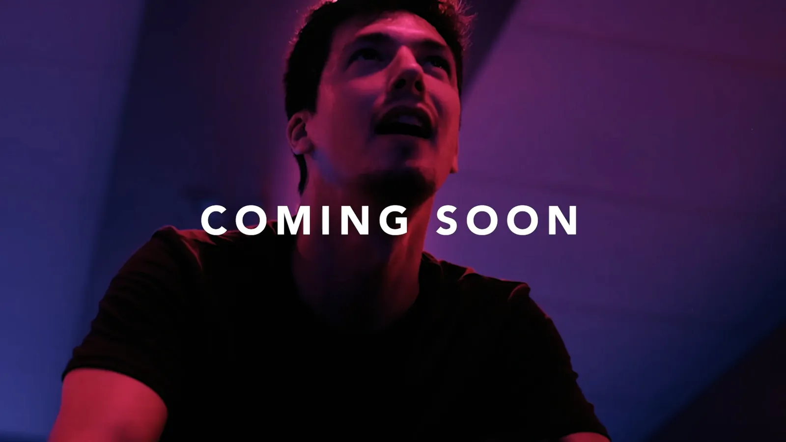 Mann im schwarzen T-Shirt blickt unter pink-blauem Licht in einem Innenraum nach oben; Schriftzug COMING SOON.
