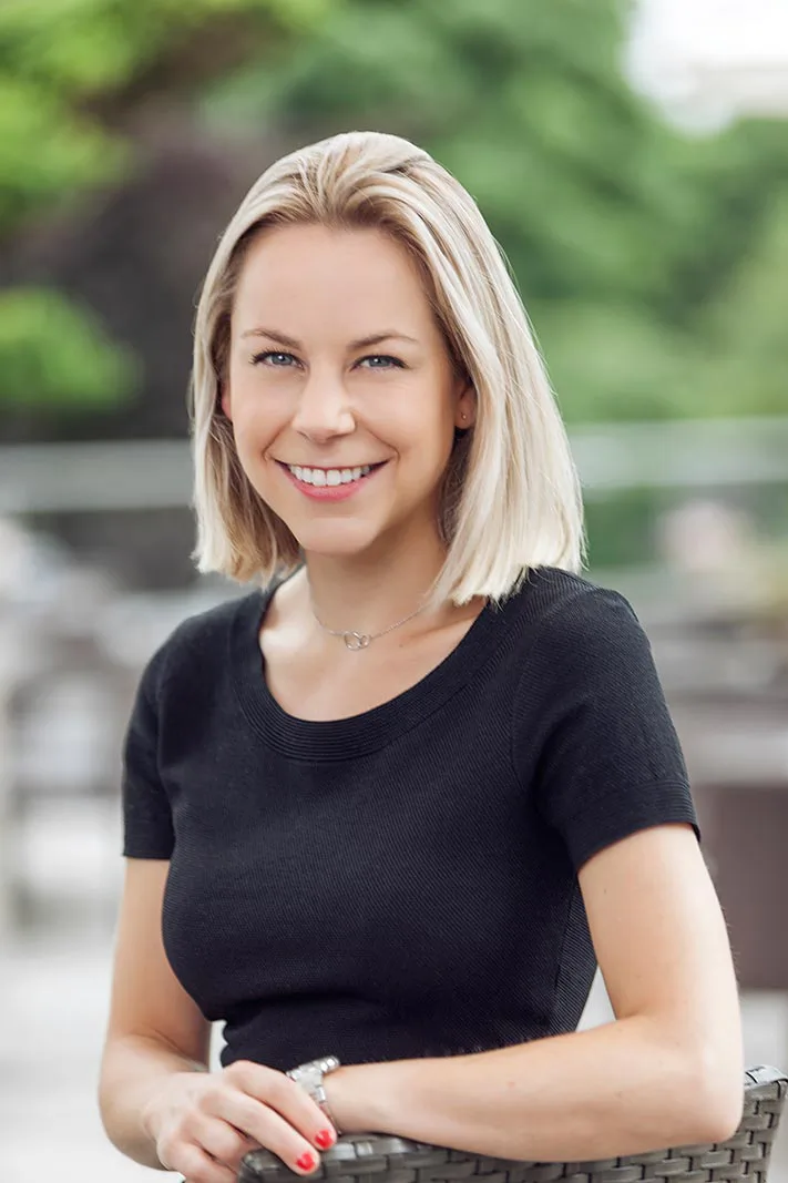 Blonde Frau im schwarzen Oberteil lächelt auf einer Terrasse bei Tageslicht, mit unscharfem Grün im Hintergrund.