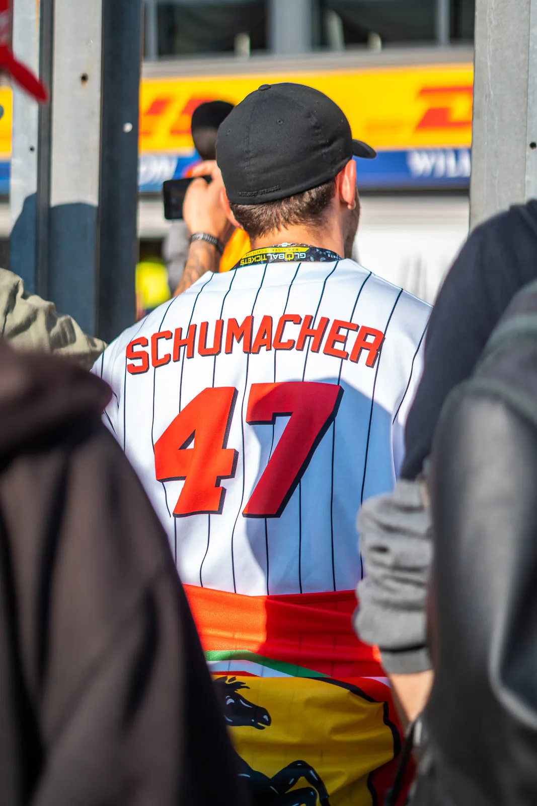 Person mit schwarzer Kappe und weißem Trikot mit roter Nummer 47 von hinten, umgeben von Menschen im Sonnenlicht.