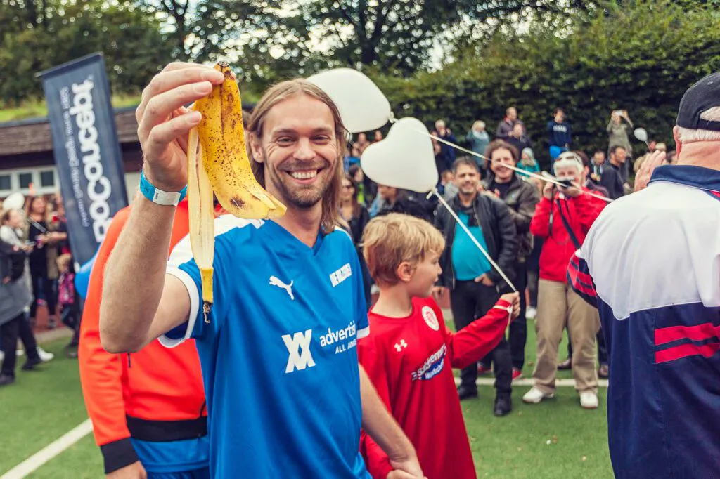 Lächelnder Mann im blauen Fußballtrikot hält eine Bananenschale hoch, umringt von Menschen auf einem Kunstrasenplatz.