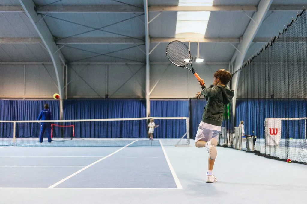 Junger Tennisspieler schlägt im Sprung einen Ball in einer lichtdurchfluteten Tennishalle mit Wilson-Banner.