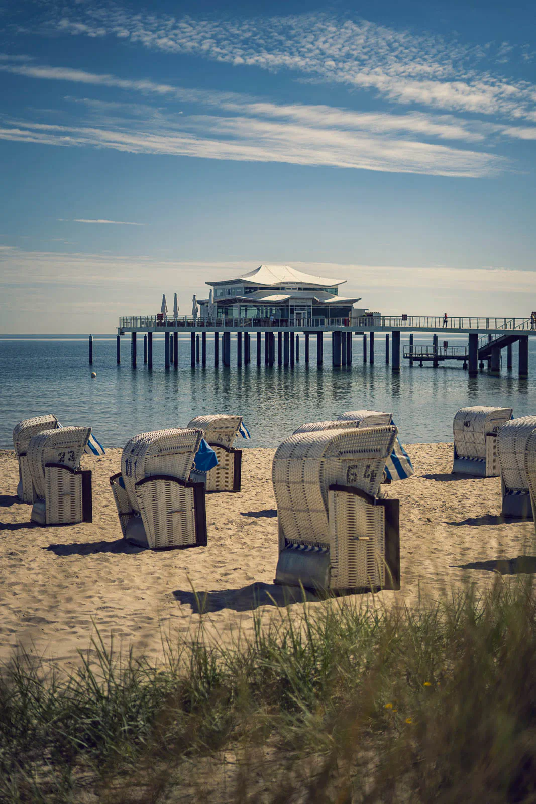 Strandkörbe im sonnigen Sand vor ruhigem Meer und einer Seebrücke mit weißem Pavillon unter blauem Himmel.