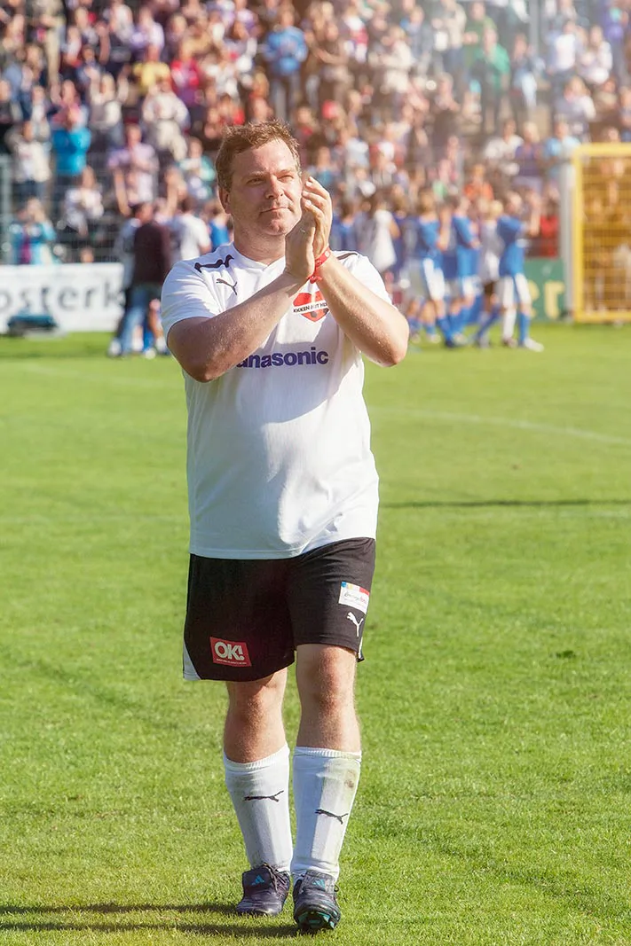 Ein Mann im weißen Fußballtrikot klatscht auf dem sonnigen Spielfeld vor einer voll besetzten Tribüne.