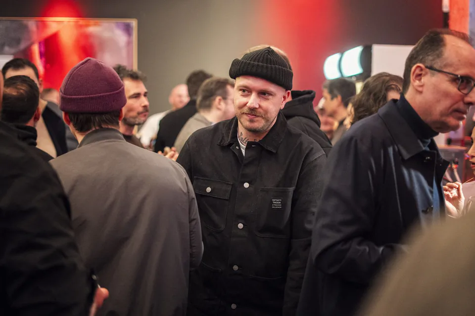 Gäste im Gespräch bei einem Event in rot beleuchtetem Innenraum, mittig ein Mann mit schwarzer Mütze und Jacke.