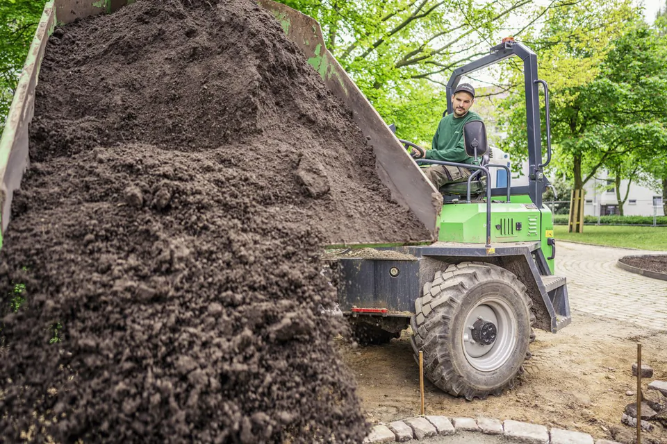 Mann am Steuer eines grünen Dumpers kippt Erde aus der Mulde auf einer Baustelle unter Bäumen.