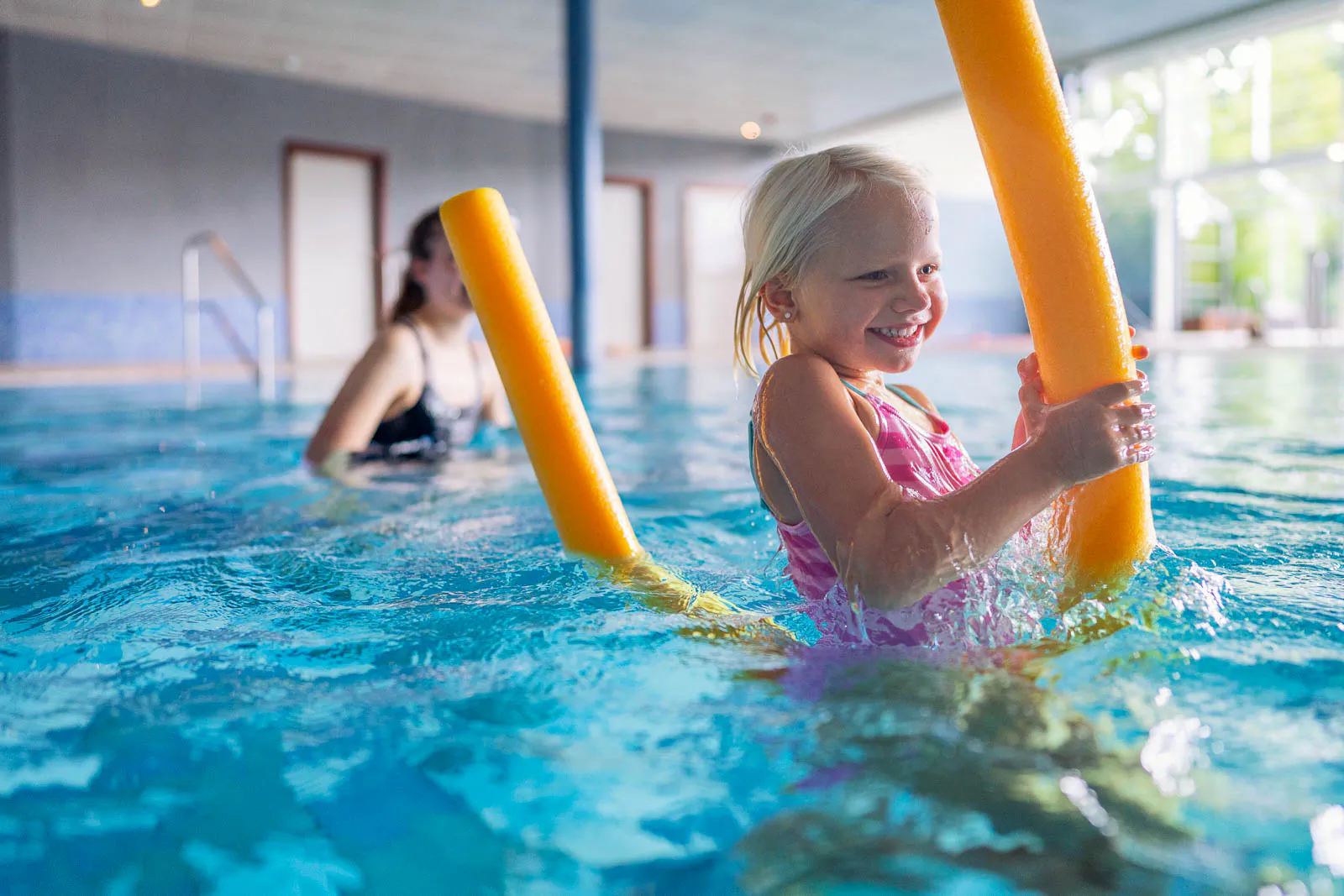 Lachendes Mädchen mit gelber Schwimmnudel im hellen Hallenbad, dahinter eine Frau im Wasser.
