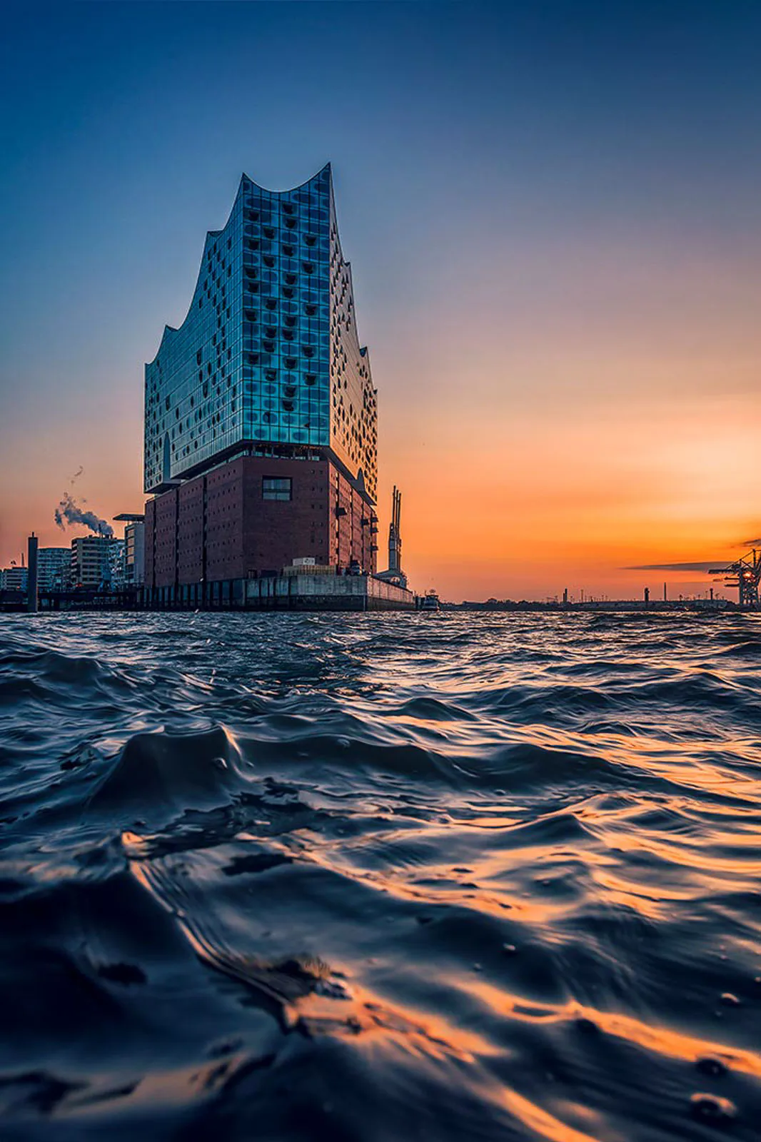 Elbphilharmonie mit gläserner Wellenfassade am Wasser, davor Wellen im orangefarbenen Abendlicht.