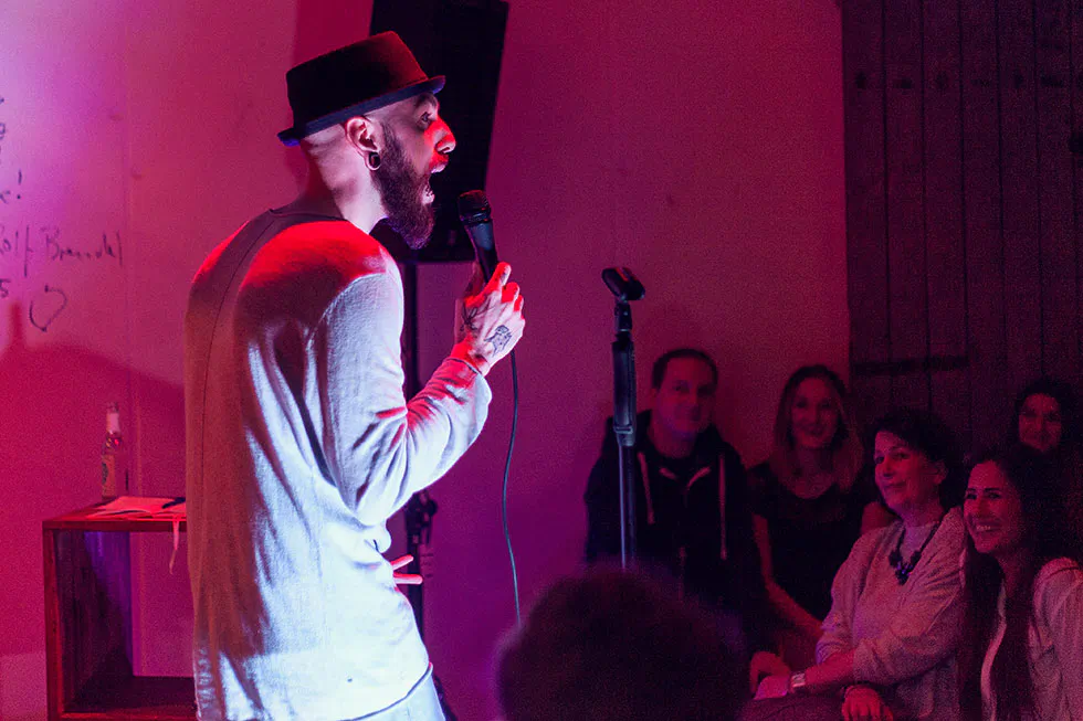 Dennis Grundt mit Hut und Mikrofon auf einer rot beleuchteten Stand-up-Bühne vor sitzendem Publikum.