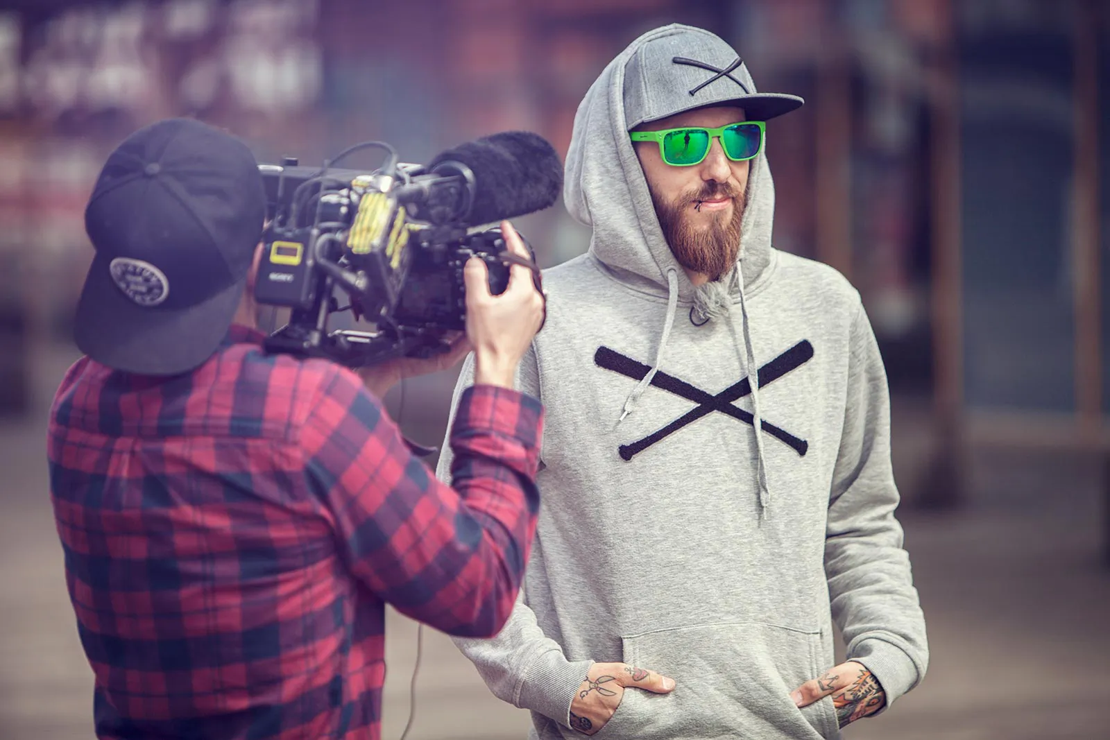 Kameramann filmt draußen einen bärtigen Mann mit grauem Kapuzenpullover, Kappe und grüner Sonnenbrille.