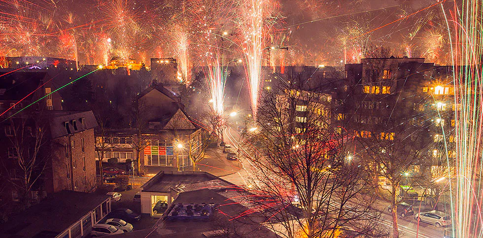 Bunte Feuerwerksspuren erhellen den Nachthimmel über Wohnhäusern, Straßen und kahlen Bäumen zum Jahreswechsel.