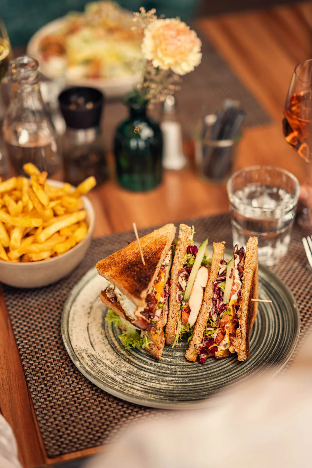 Belegte Sandwichdreiecke auf einem Keramikteller, daneben Pommes und Wasserglas auf einem warm beleuchteten Holztisch.