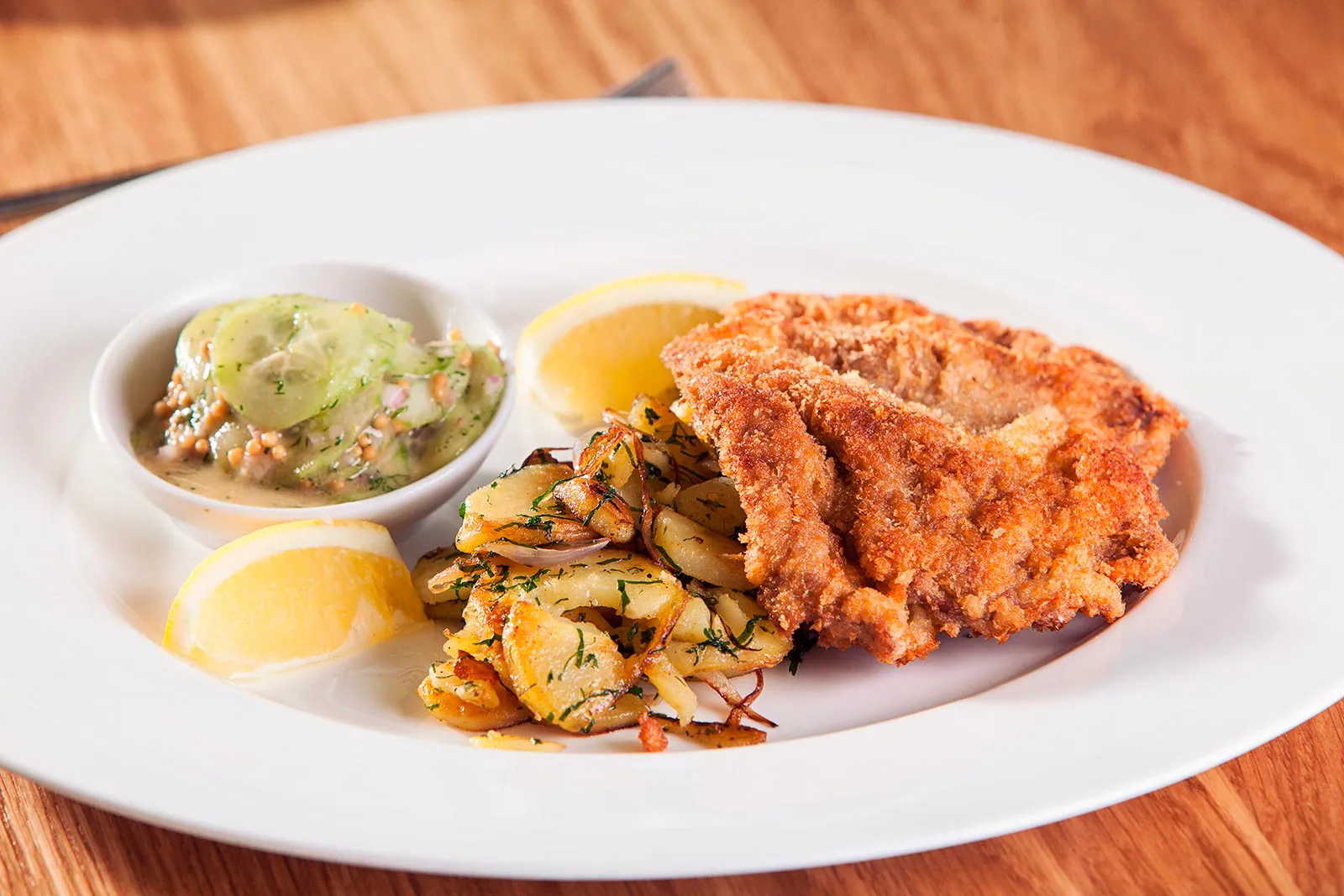 Goldbraunes Schnitzel mit Bratkartoffeln, Gurkensalat und Zitronenspalten auf weißem Teller auf einem Holztisch.