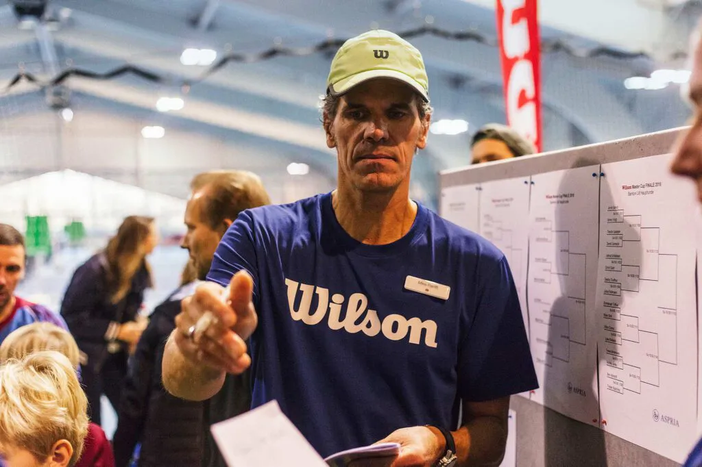 Mann in blauem Wilson-Shirt und Kappe gestikuliert neben einem Turnierplan in einer beleuchteten Sporthalle.