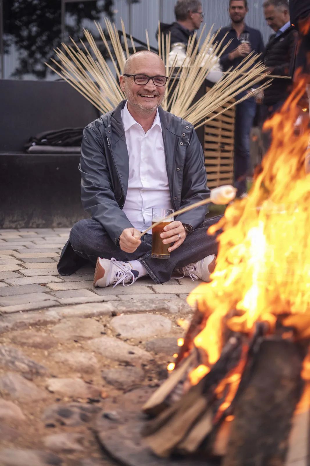 Lachender Mann mit Brille sitzt vor einem Lagerfeuer und hält einen Stock mit Marshmallow über die Flammen.
