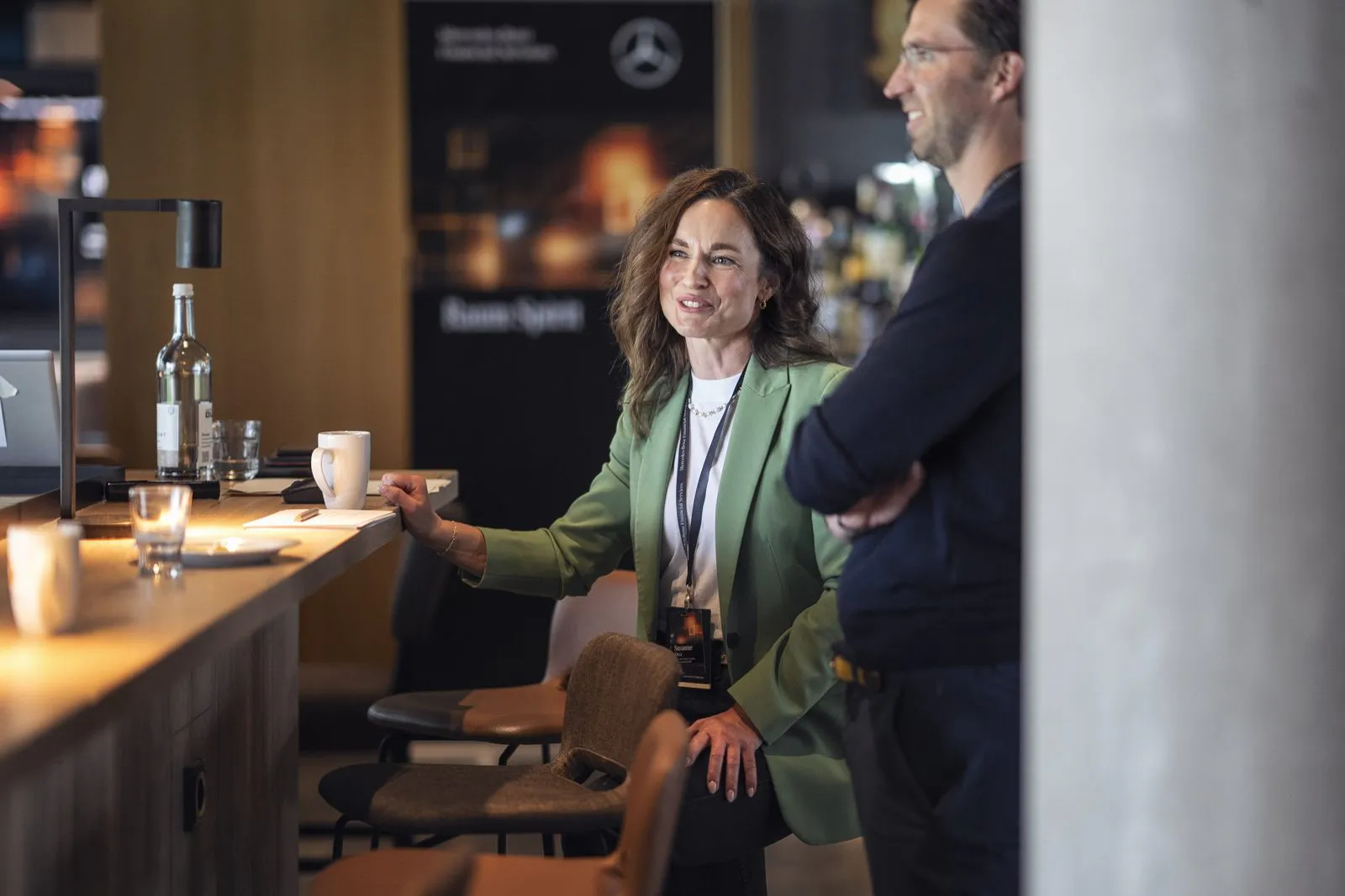 Frau im grünen Blazer und Mann an einer warm beleuchteten Bar, Mercedes-Benz-Eventplakat im Hintergrund.