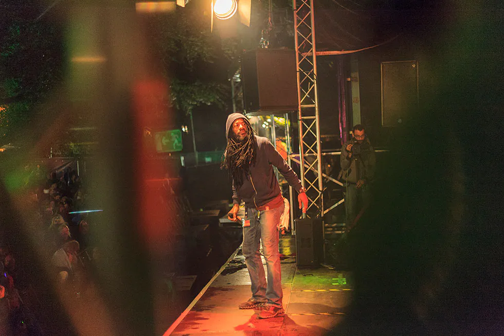 Sänger mit Dreadlocks und Kapuze hält ein Mikrofon auf der Bühne, beleuchtet von warmem gelb-rotem Licht.