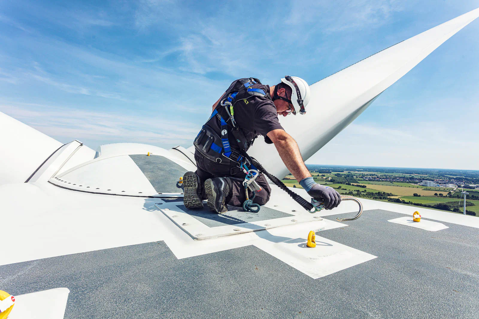 Techniker mit Helm und Sicherungsgurt kniet bei Sonnenschein auf einer Windkraftanlage neben einem Rotorblatt.
