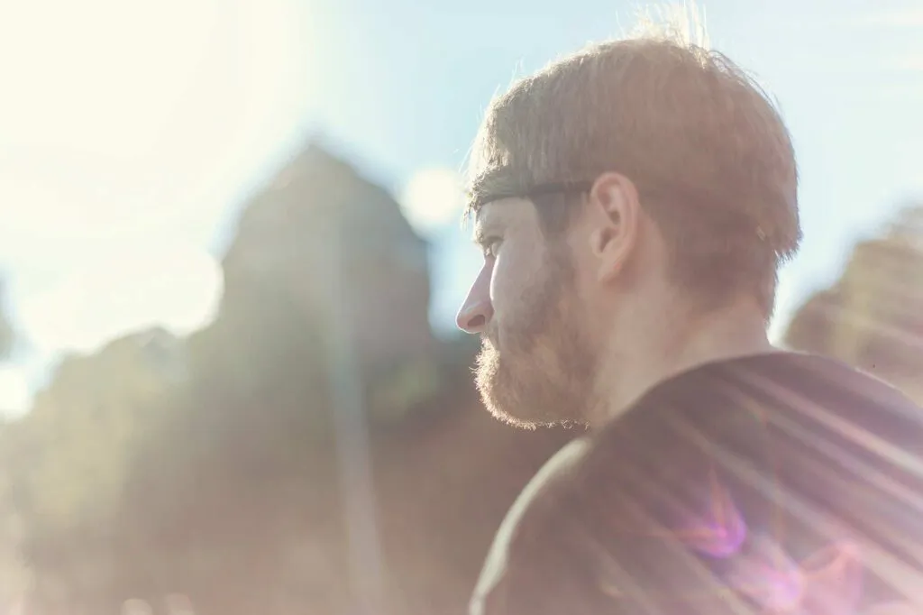 Bärtiger Mann im Profil, draußen im starken Gegenlicht vor unscharfen Gebäuden und Bäumen.