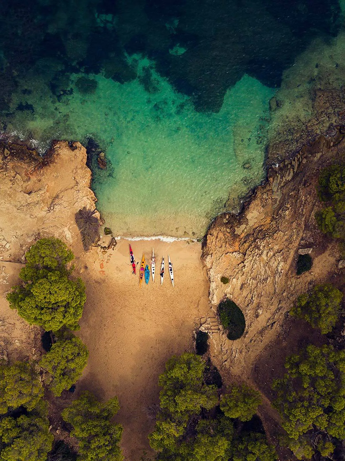 Drohnenaufnahme einer felsigen Bucht mit türkisgrünem Wasser, schmalen Kajaks am Sandstrand und grünen Bäumen.