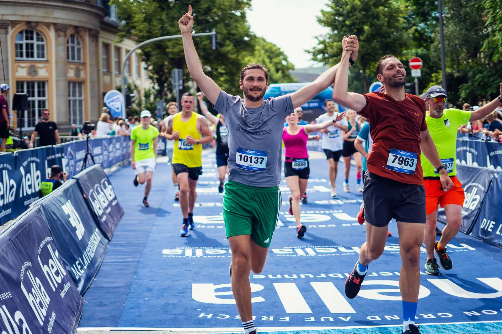 Zwei Läufer jubeln Hand in Hand auf der blauen Zielgeraden des Hella Halbmarathons, weitere Läufer folgen.