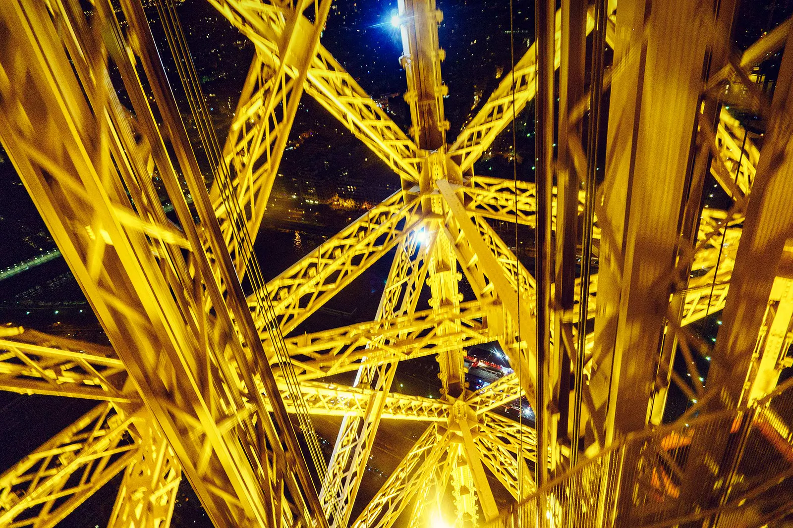 Blick durch die goldgelb beleuchteten Stahlstreben des Eiffelturms bei Nacht, mit Lichtern der Stadt im Hintergrund.