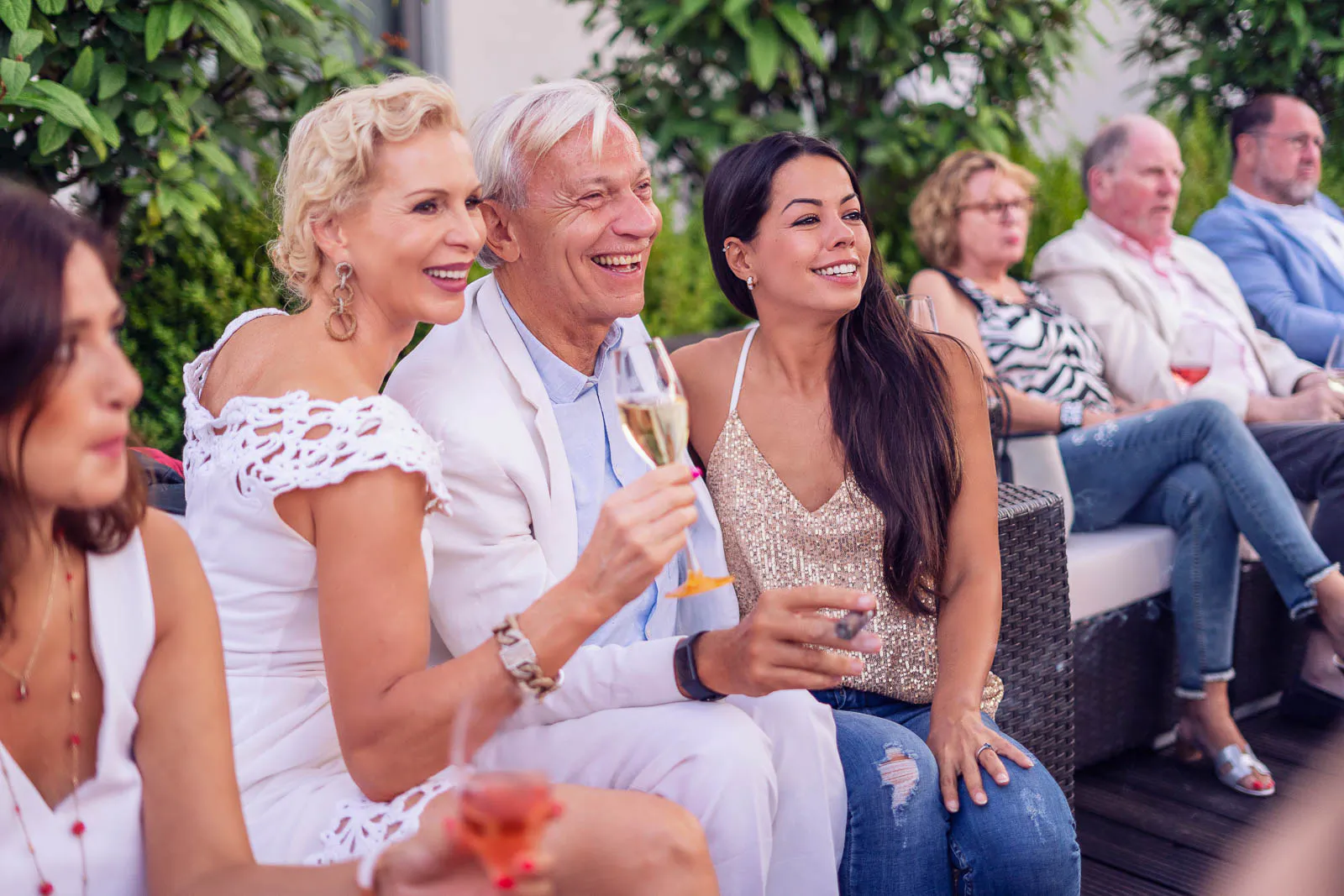 Drei lächelnde Partygäste mit Sekt- und Wasserglas sitzen bei Tageslicht auf einer Terrasse vor grünen Pflanzen.