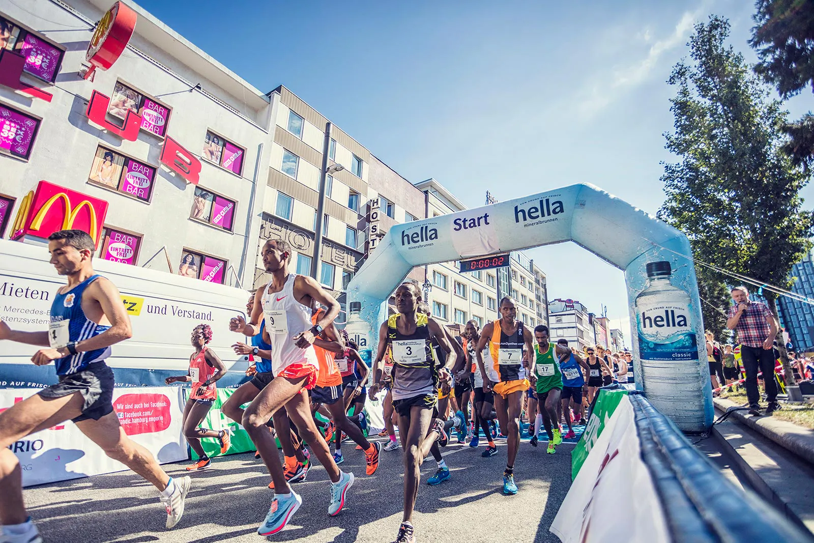 Läufer starten beim Hella Halbmarathon unter einem weißen Startbogen auf einer sonnigen, abgesperrten Straße.