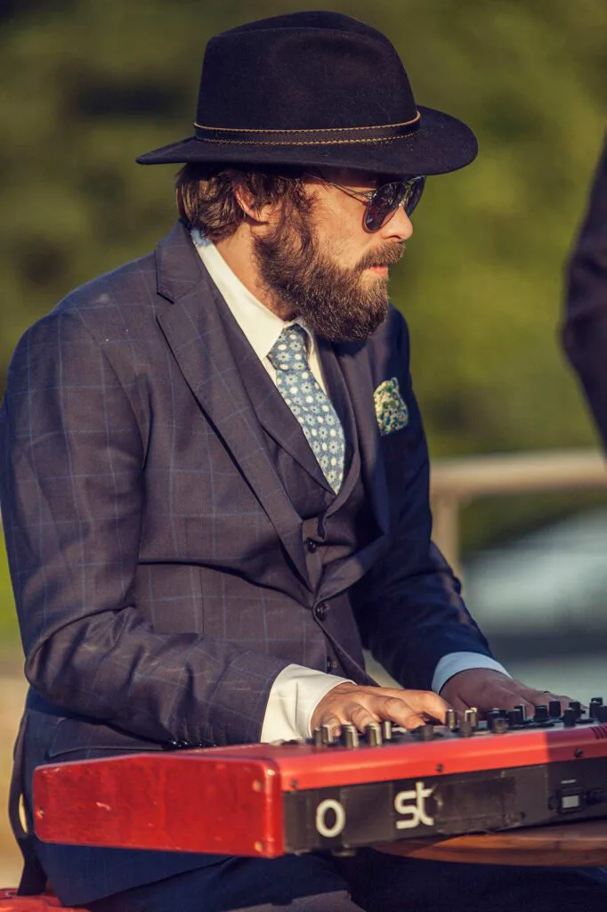 Bärtiger Musiker mit Hut, Sonnenbrille und Anzug spielt bei warmem Licht draußen an einem roten Keyboard.