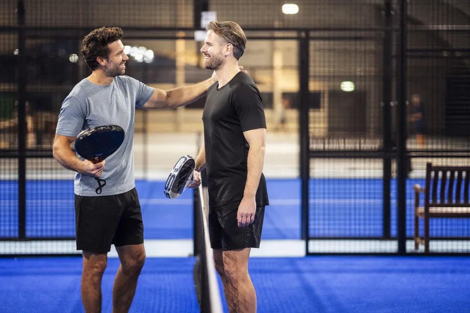 Zwei lächelnde Männer mit Padelschlägern stehen am Netz eines blauen Indoor-Platzes im warmen Hallenlicht.