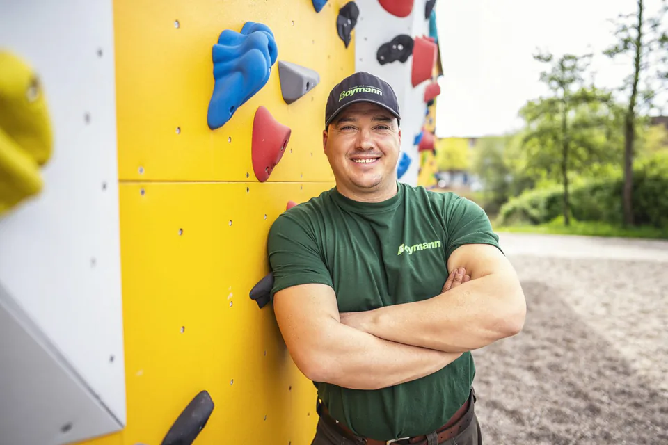 Lächelnder Mann in grünem Boymann-Shirt und Kappe steht mit verschränkten Armen an einer gelben Kletterwand im Freien.