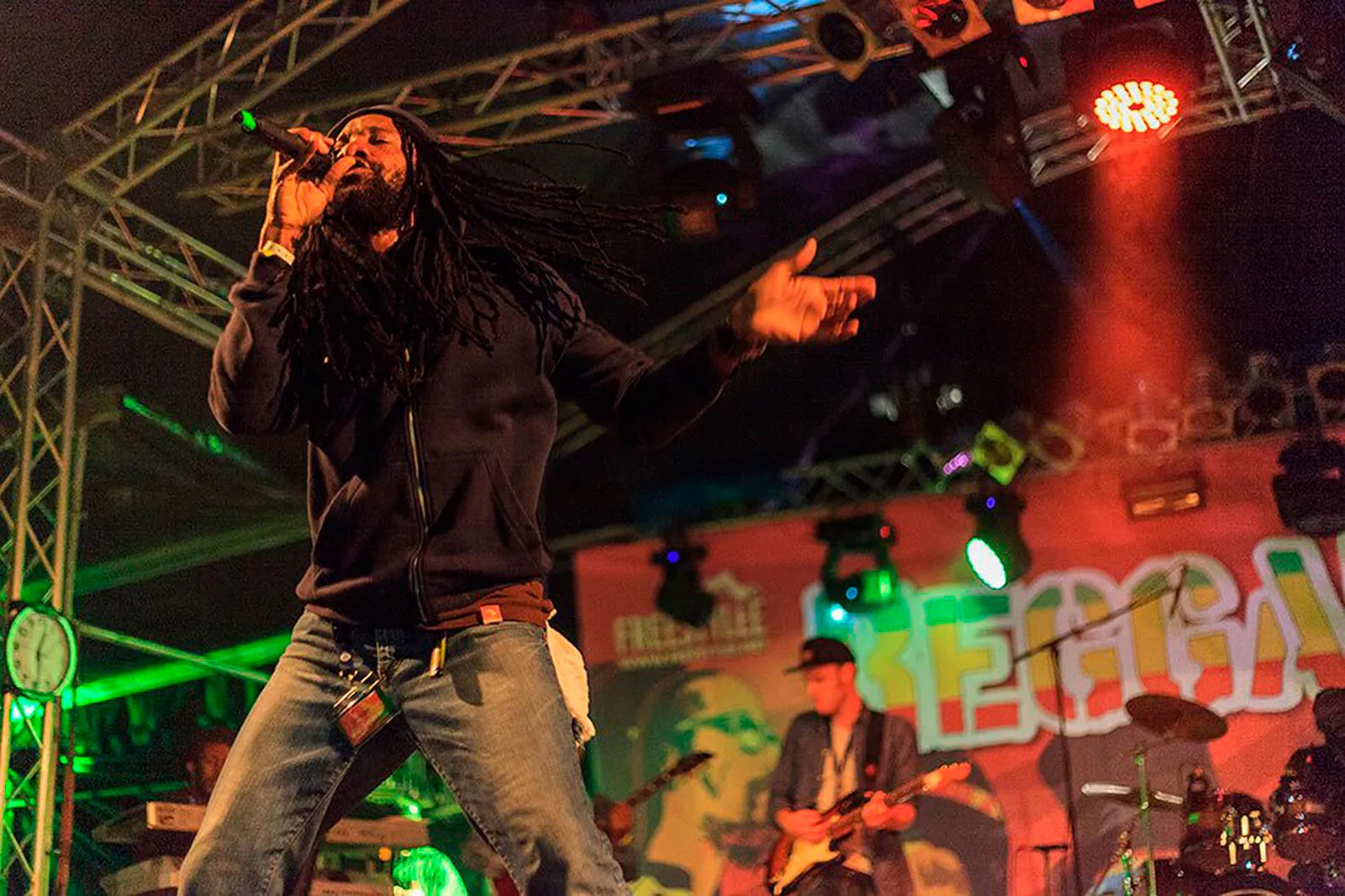 Sänger mit langen Dreadlocks singt beim Reggae Jam ins Mikrofon, hinter ihm Musiker im roten und grünen Bühnenlicht.